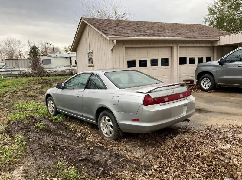 Silver Honda coupe parked on muddy ground near a garage.