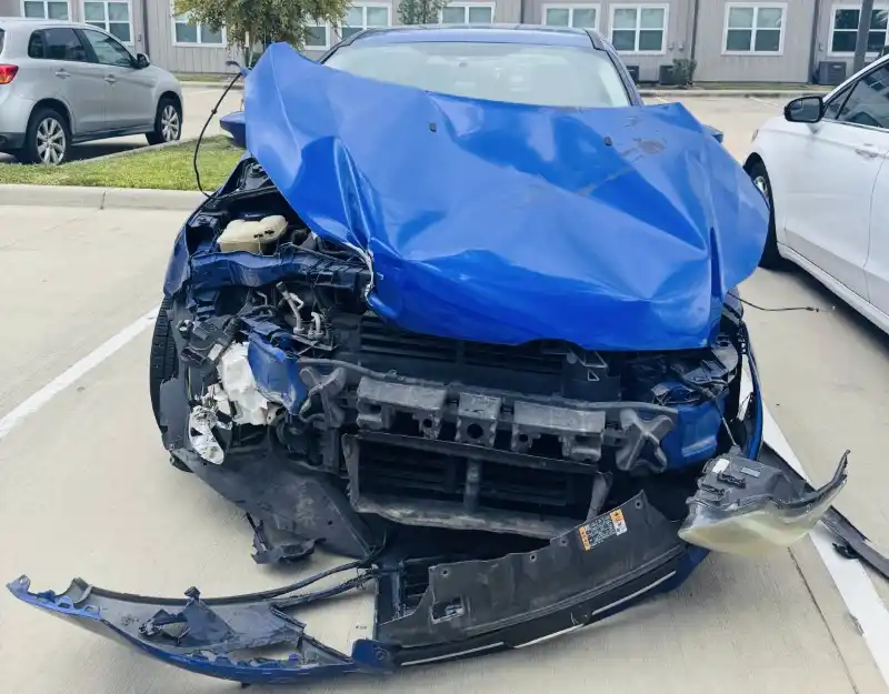 Blue wrecked car with severe front-end damage parked in Fresno, TX.